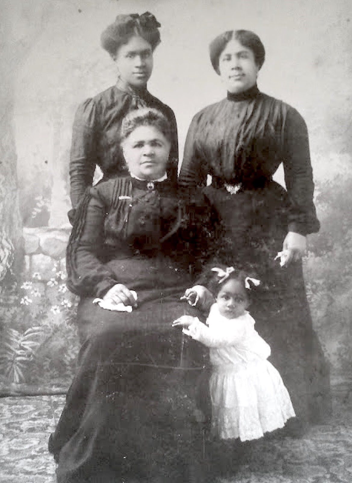 Four women in period dress, one sitting and two standing pose for a photograph with a little girl in a white dress stands, holding onto the leg of the sitting woman.